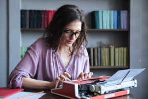 Brunette mature woman journalist in eyeglasses typing on typewriter indoors